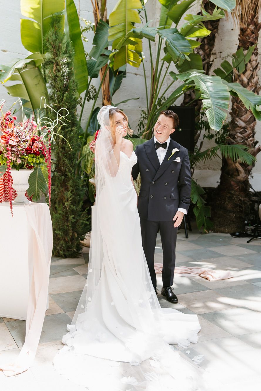 Bride in white gown and veil laughs with groom in black tuxedo outdoors among tropical greenery at Masia Victoria