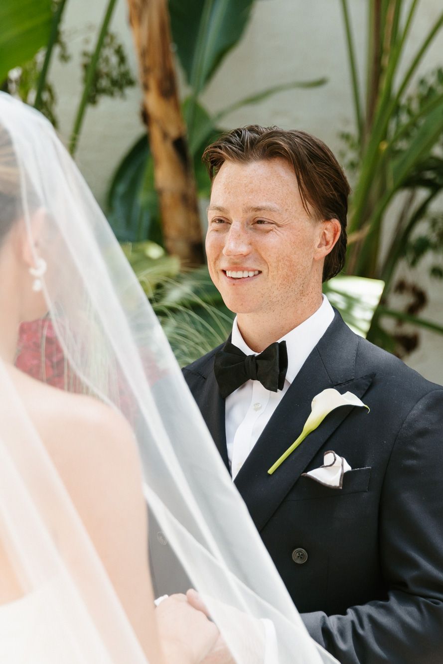 Smiling groom in black tuxedo with calla lily boutonniere gazing at bride during outdoor ceremony.