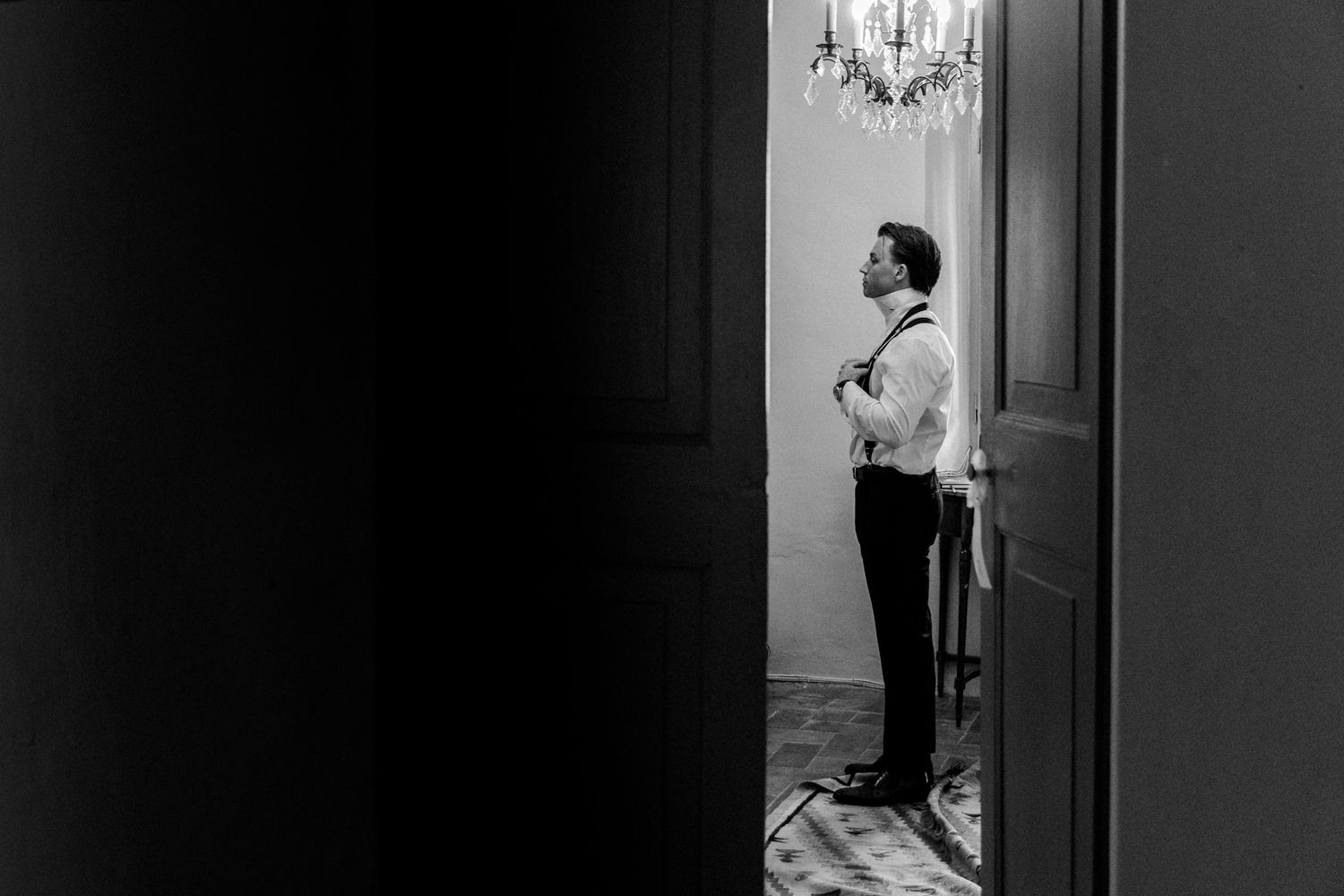 Black and white photo of a groom adjusting his tie in a doorway beneath an elegant chandelier.