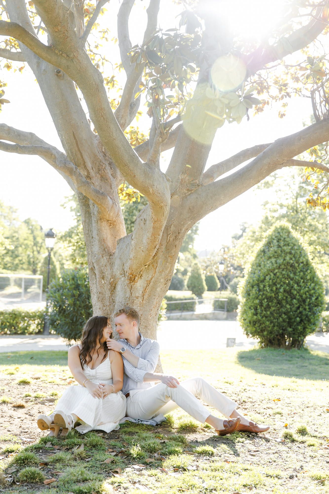 Couple sits together under large tree in sunny garden, sharing an intimate moment in white outfits.