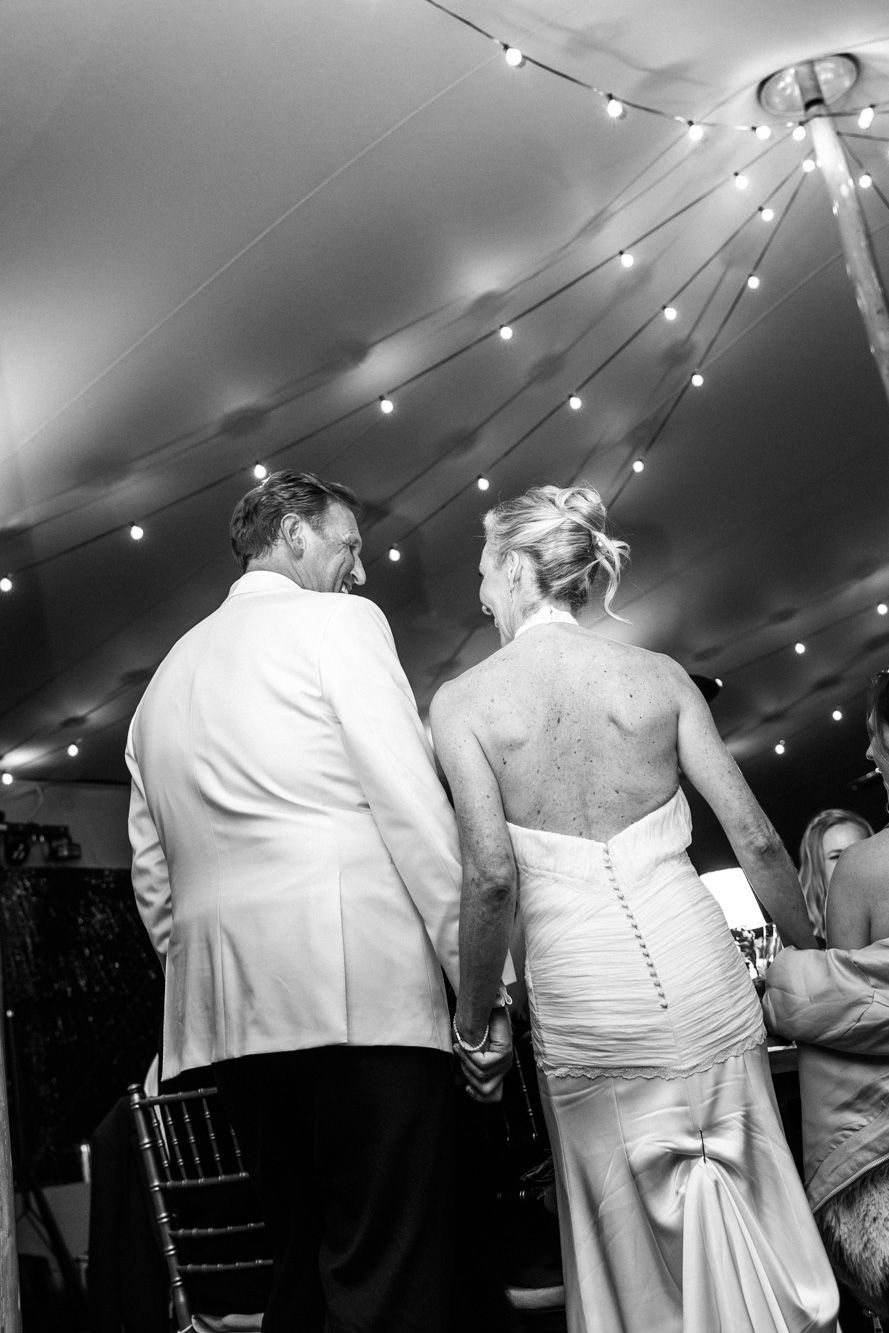 Black and white photo of a bride and groom holding hands and smiling at each other under a tent with string lights at a wedding reception.