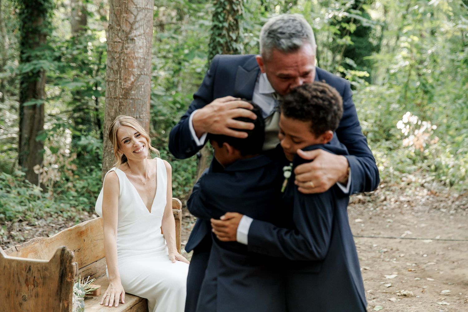 Groom hugging children during outdoor woodland wedding ceremony while bride in white dress smiles warmly nearby.