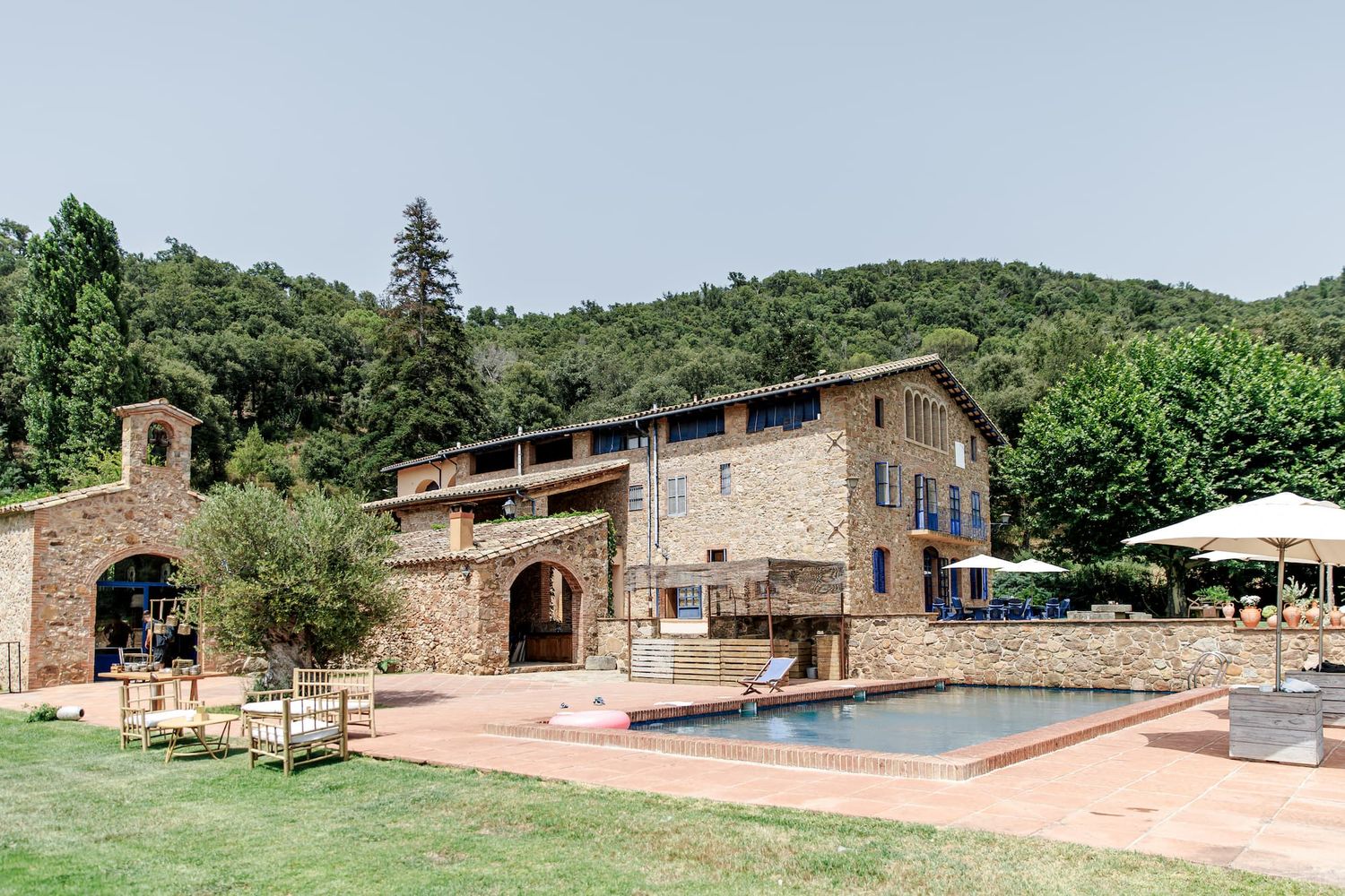 Rustic stone villa with pool surrounded by lush green hills and trees under a clear blue sky.