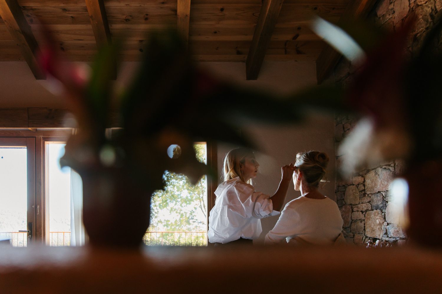 Two women in white robes get ready together in a rustic stone room with wooden beams, framed through blurred flowers in the foreground.
