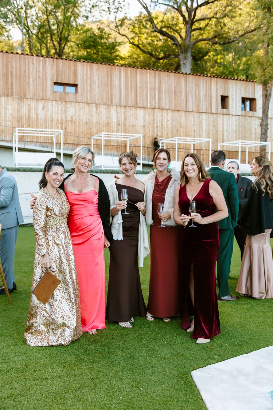 Five women in elegant evening gowns hold champagne glasses and smile together on a green lawn at an outdoor wedding reception.