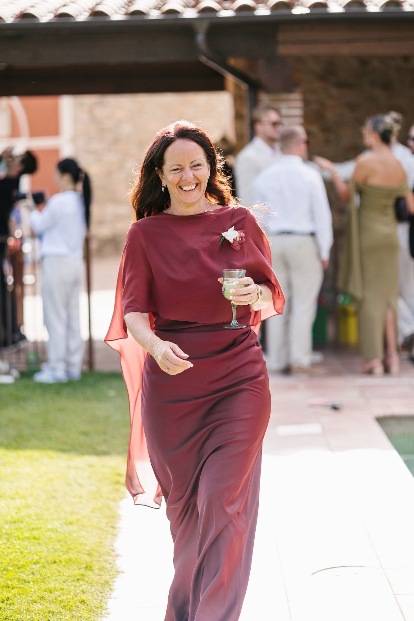 Woman in elegant burgundy gown laughing and holding a champagne glass at an outdoor wedding reception.