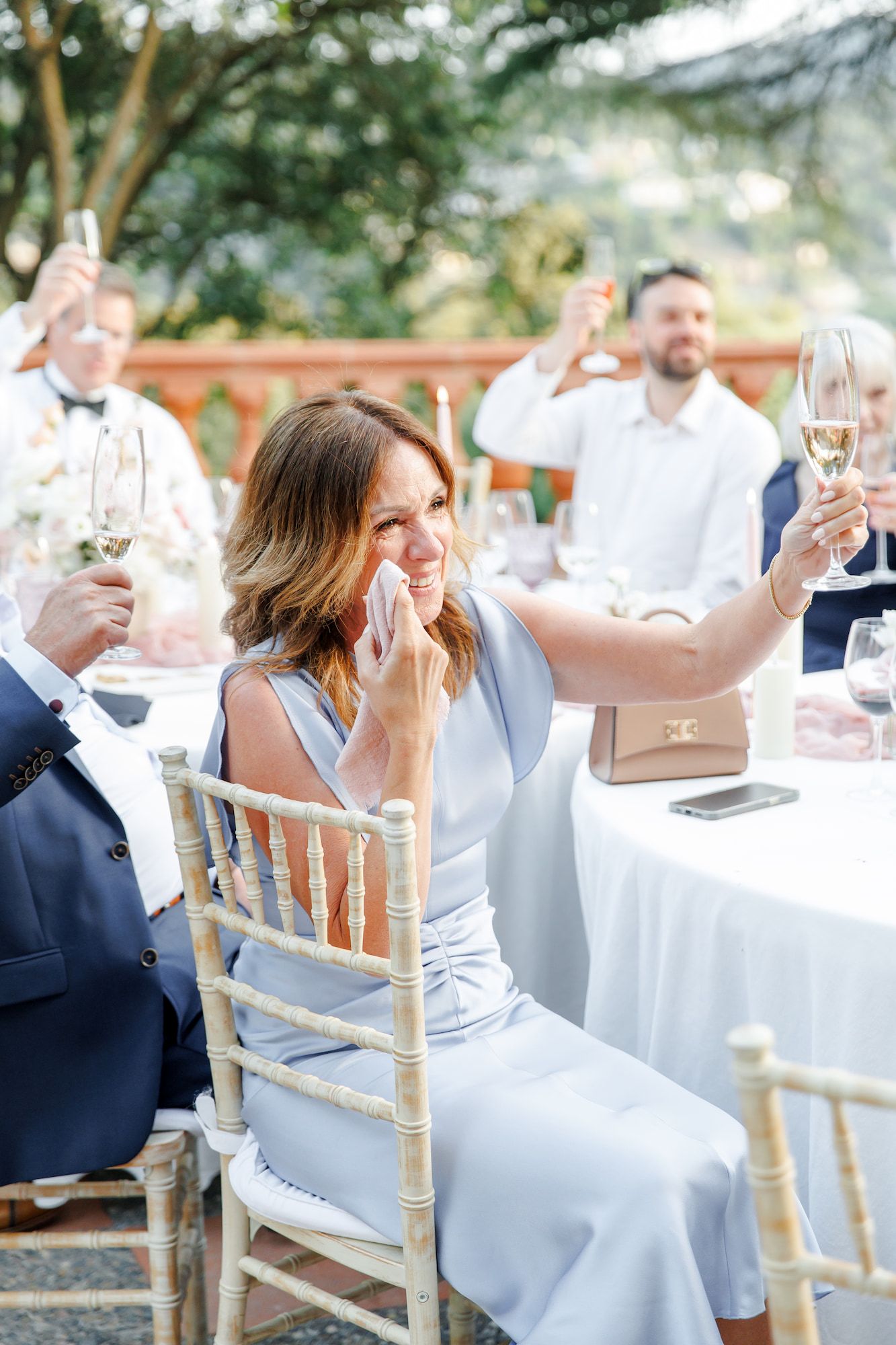 Wedding guest in blue dress raises champagne glass during toast, wiping tears at outdoor reception.