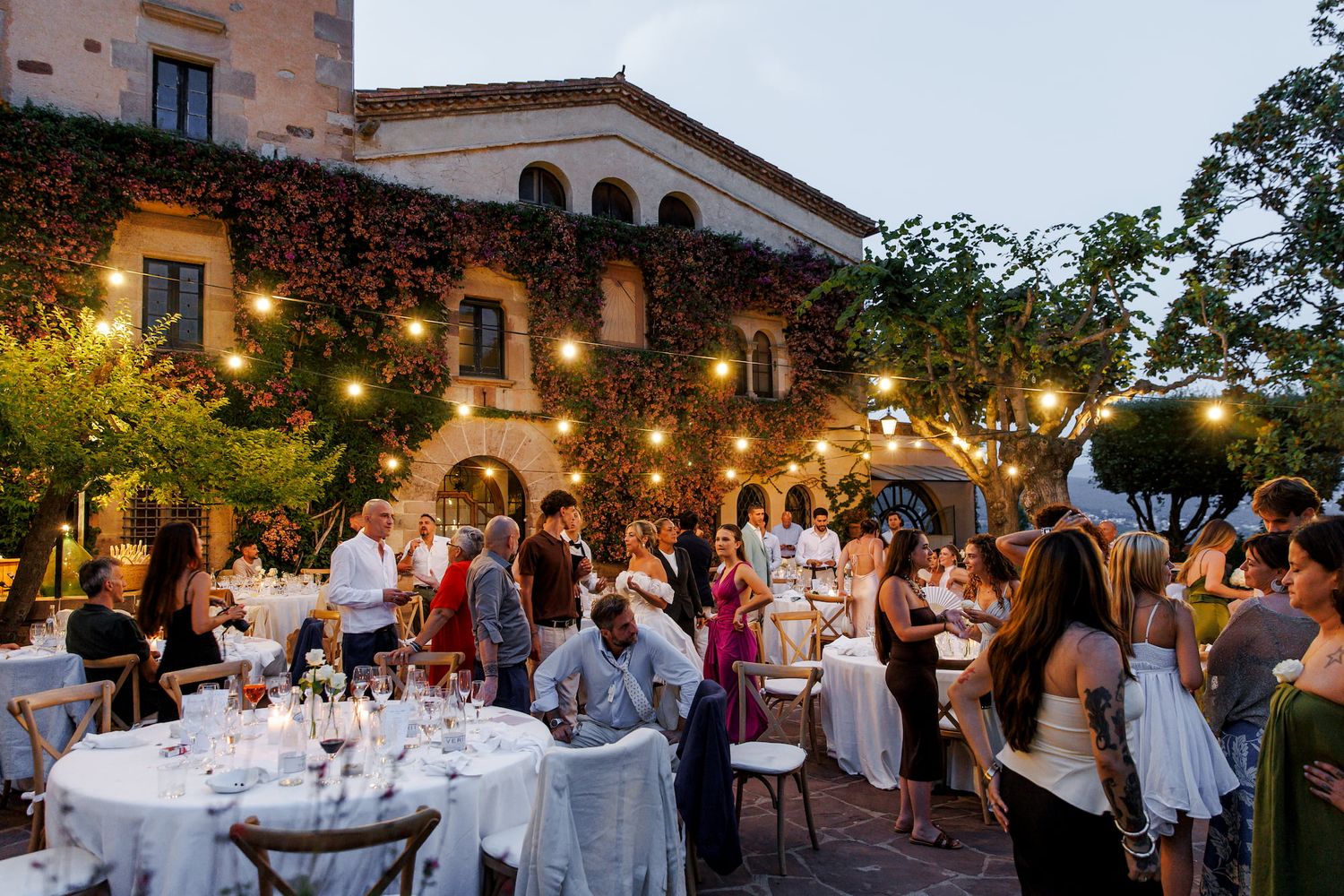 Elegant outdoor wedding reception at an ivy-covered villa with string lights and guests dining al fresco at dusk.