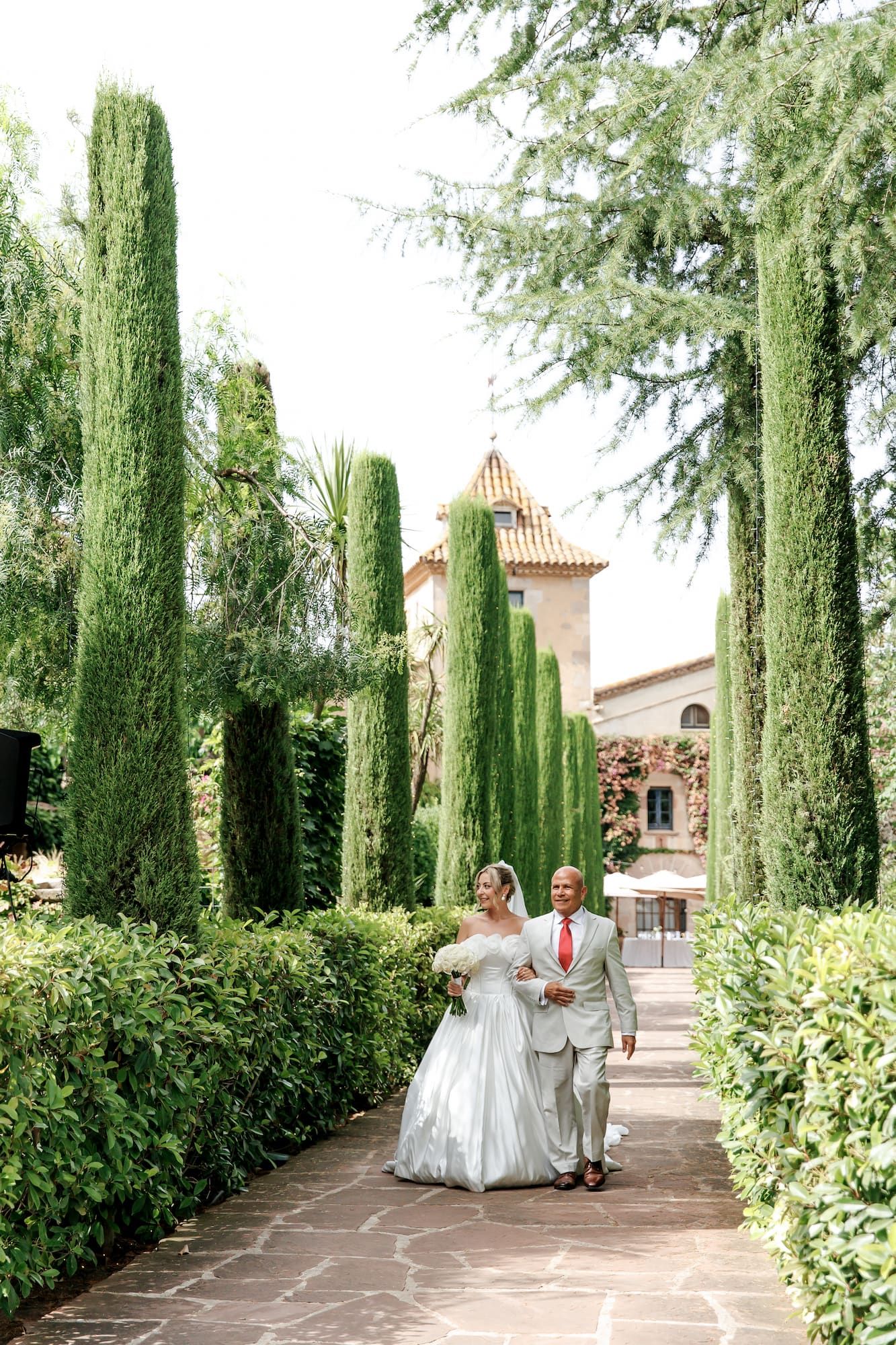 A bride and groom walk hand-in-hand along a garden path lined with tall cypress trees and a historic villa backdrop.