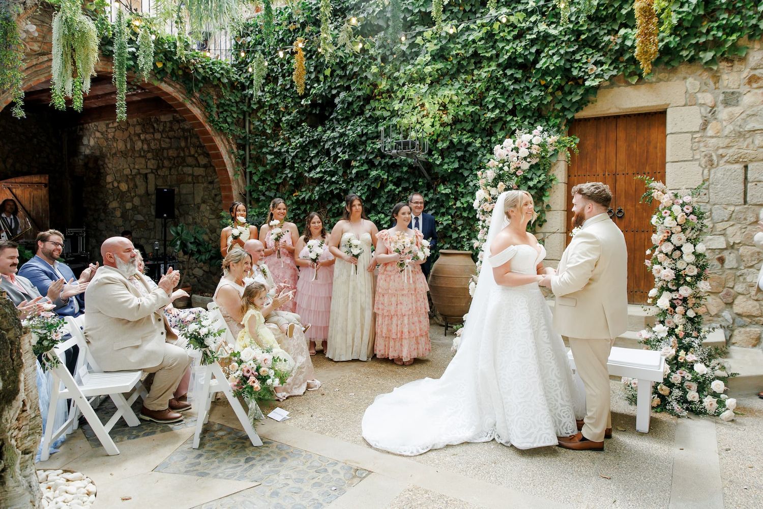 Outdoor wedding ceremony in a stone courtyard with bridesmaids, flower girls, and floral arch decorations.