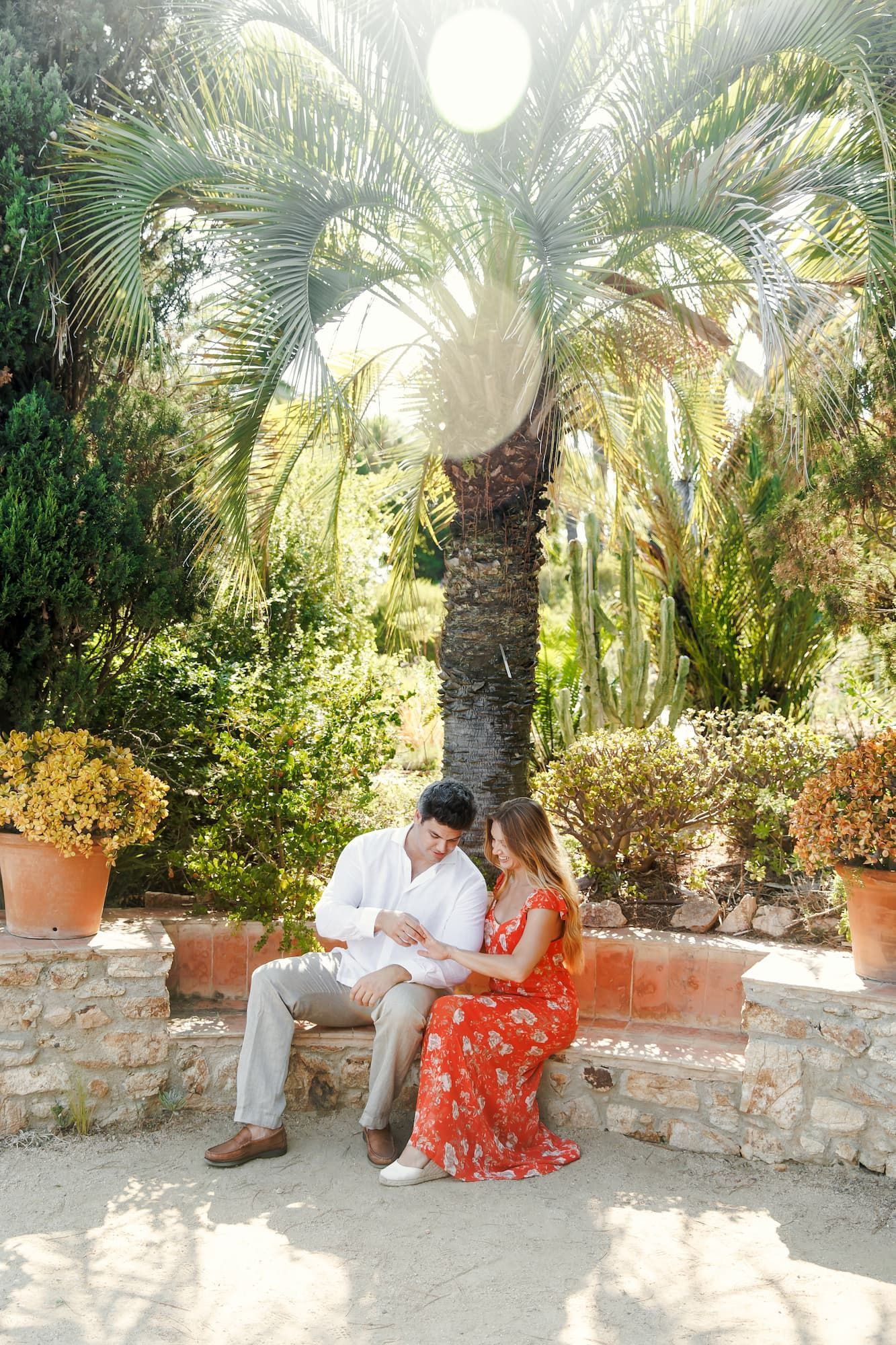 Couple sitting together on a stone bench in a lush tropical garden with palm trees, man in white shirt, woman in red floral dress.