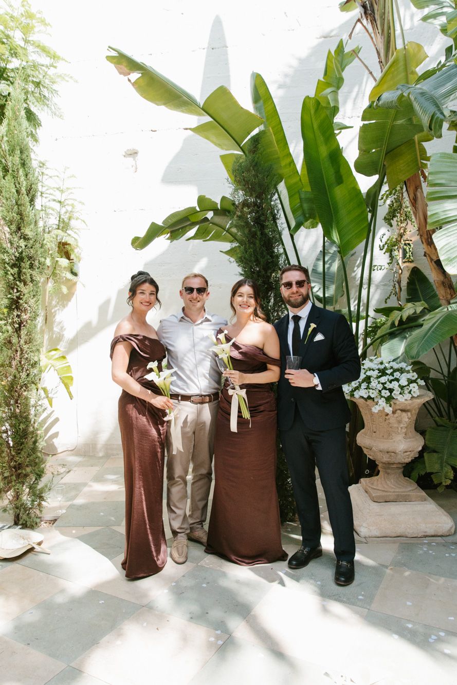 Two bridesmaids in brown satin gowns pose with groomsmen in a sunlit garden courtyard with tropical plants.