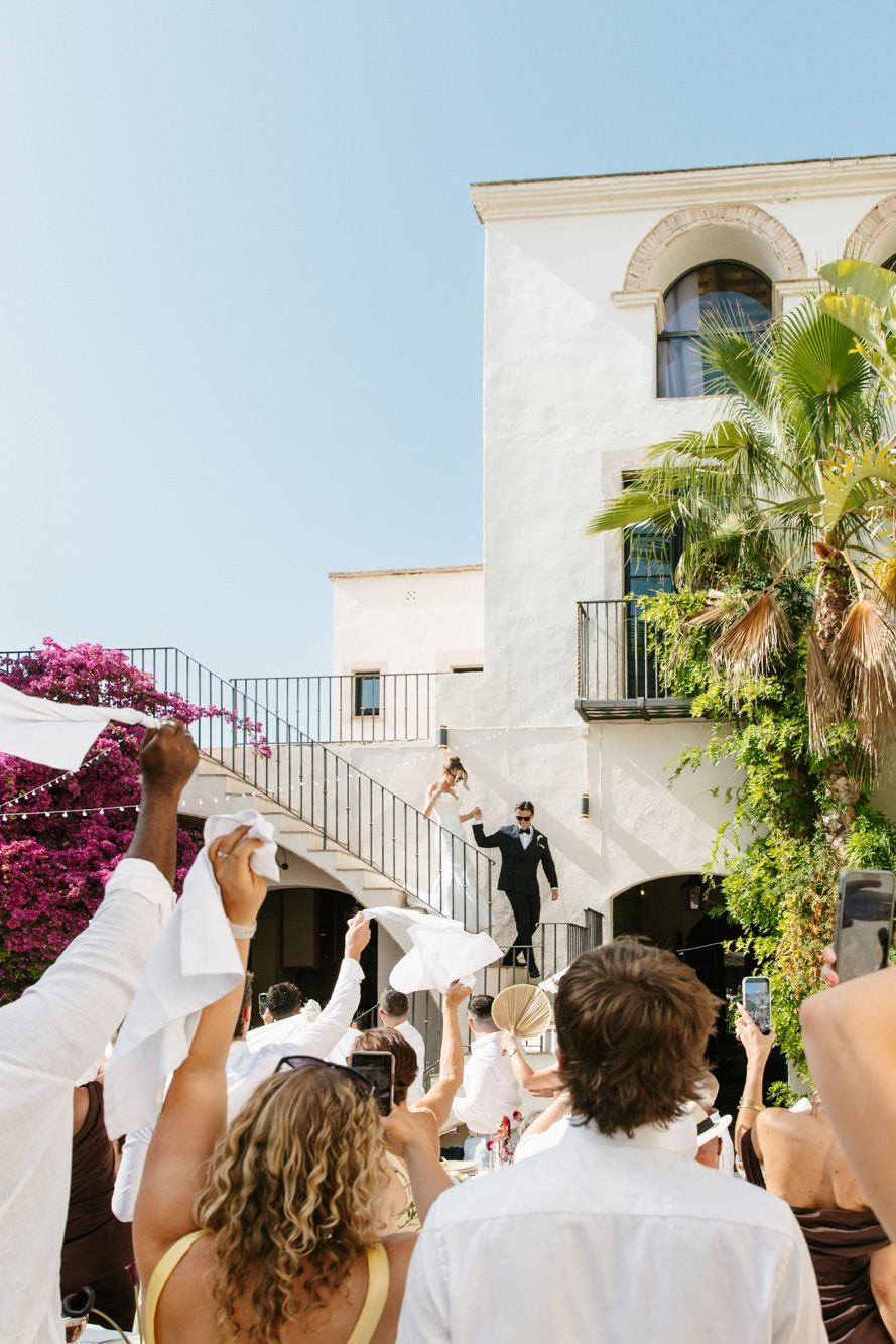 Guests wave white handkerchiefs celebrating a wedding couple descending outdoor stairs at a Mediterranean villa.