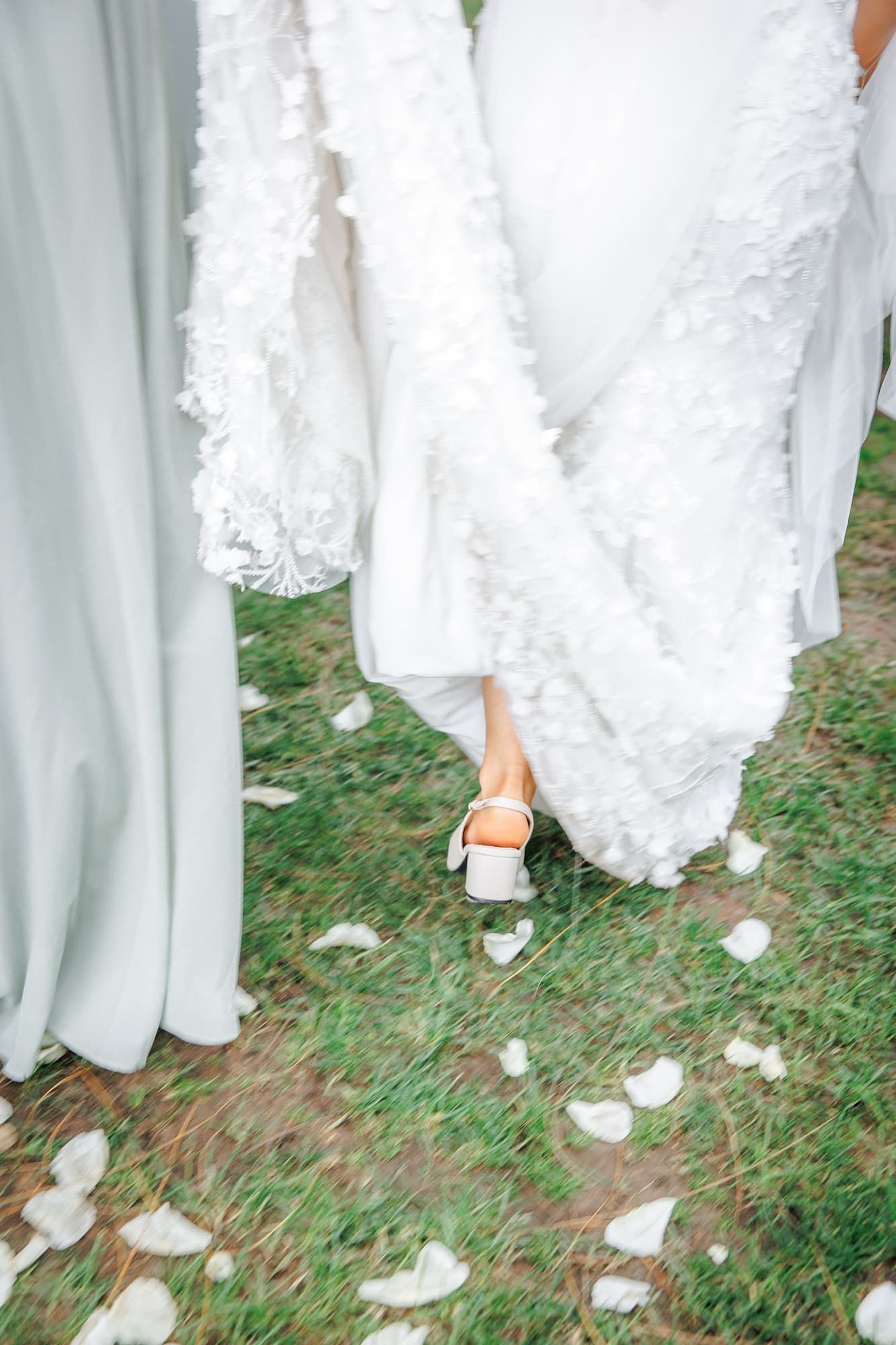 Bride in white textured gown and heels walking on grass scattered with flower petals beside bridesmaid.