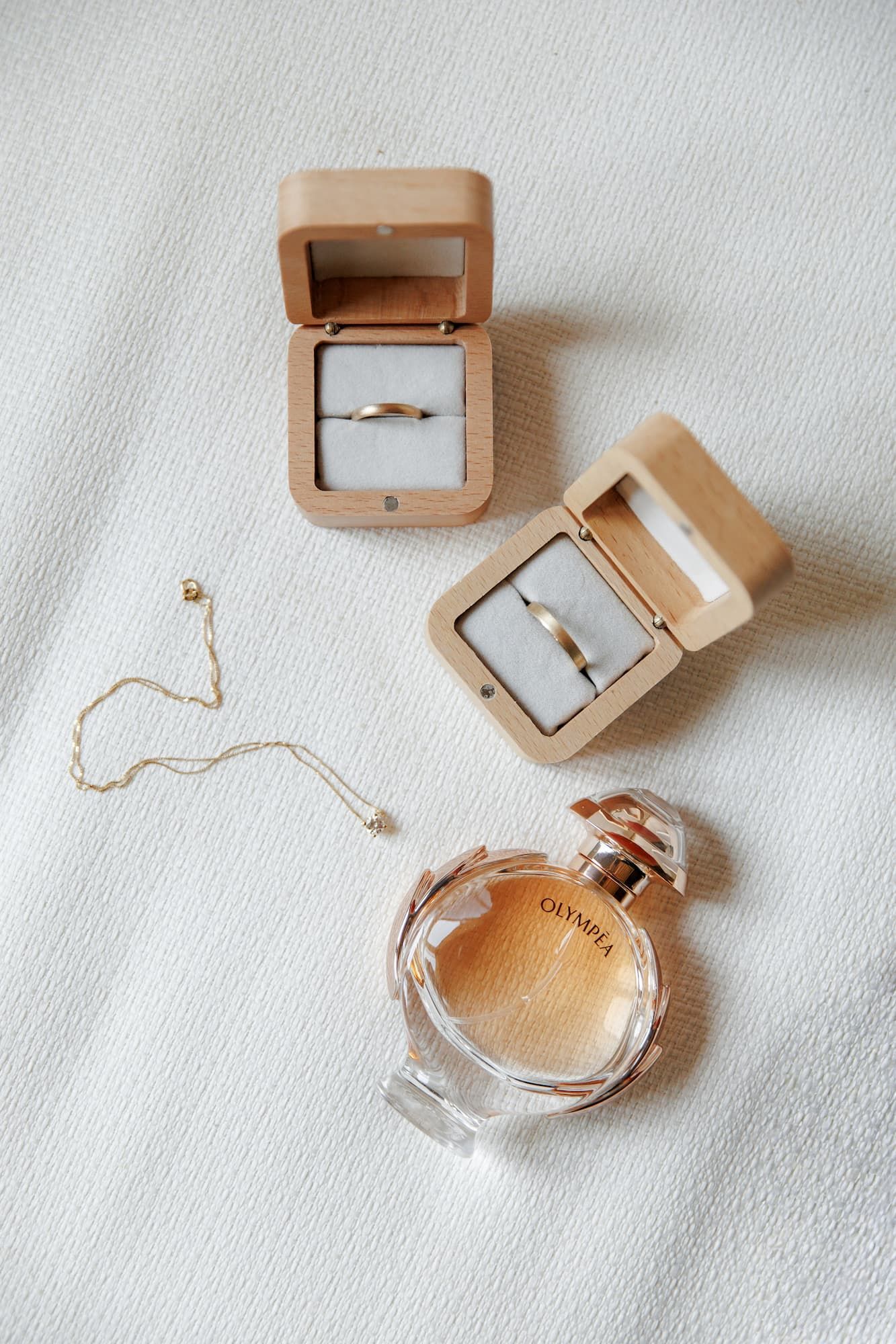 Elegant wedding flat lay with gold rings in wooden boxes, delicate gold necklace, and Olympea perfume bottle on white fabric.