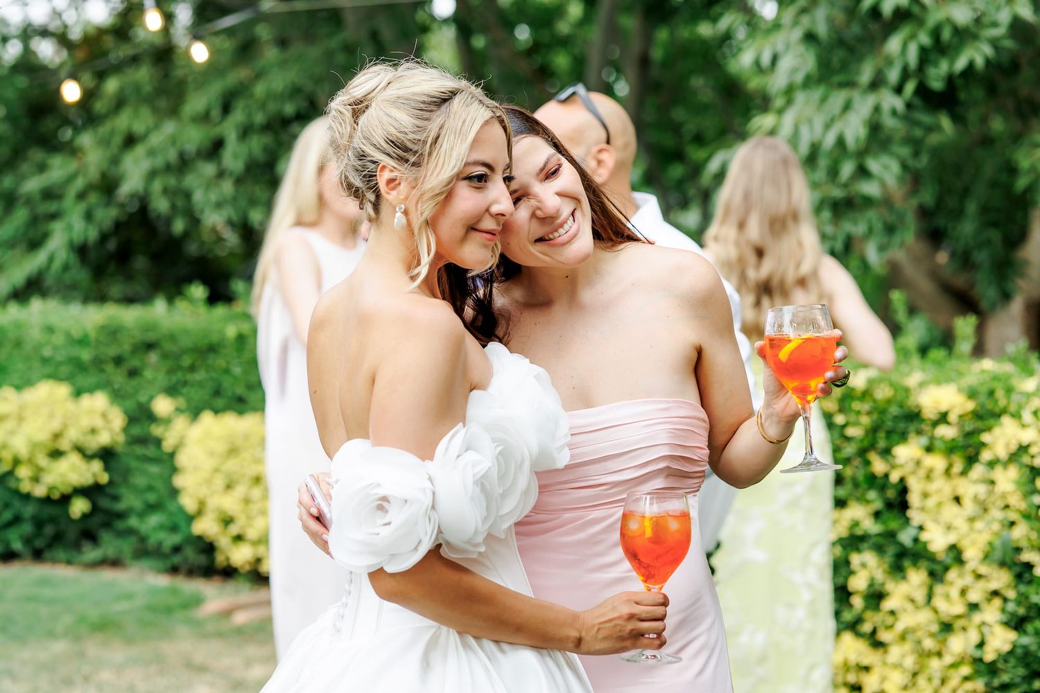 Two women in elegant dresses embrace outdoors at a garden party, one holding a colorful cocktail.