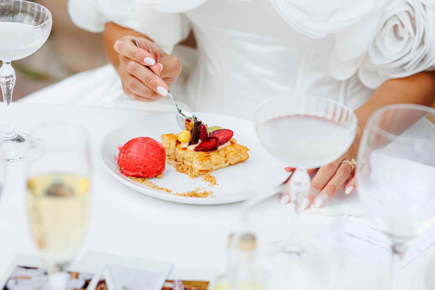 Bride in white dress eating a dessert plate with strawberries and red sorbet at a wedding reception.
