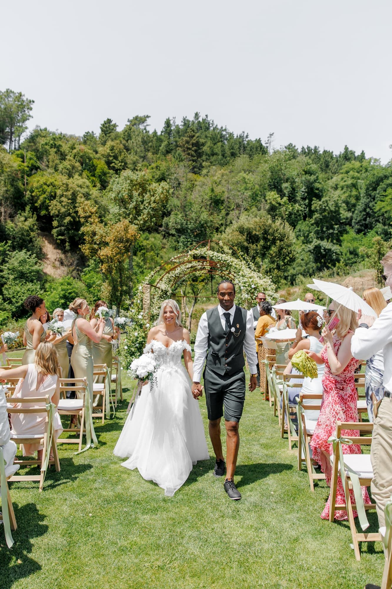 Bride and groom walk down the wedding aisle hand in hand, guests cheering, floral arch backdrop, lush green hillside.