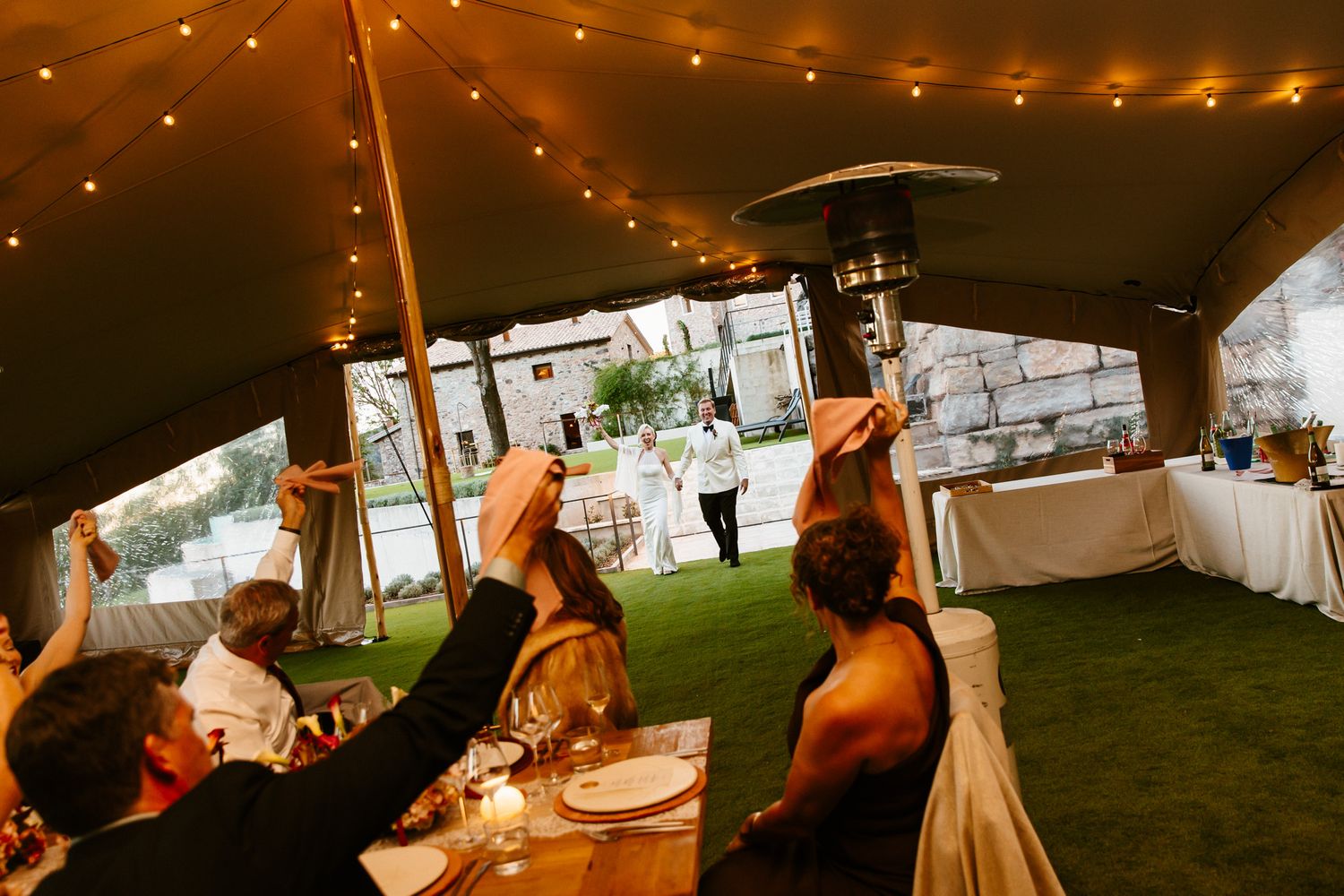 Wedding guests raise their hands and cheer under a lantern-lit tent while a couple stands on the lawn near a patio heater.