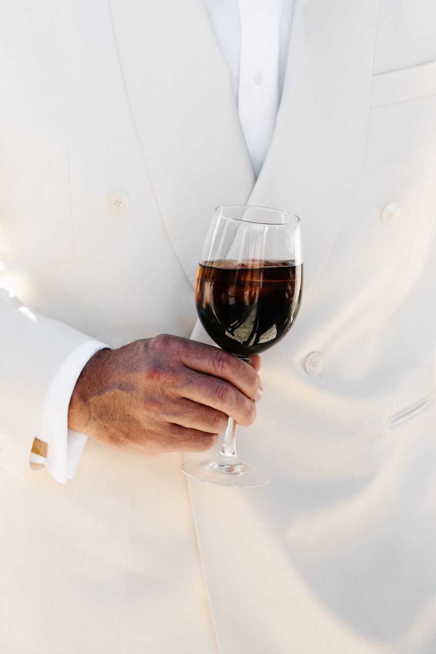 A man in a white double-breasted jacket and cufflinks holds a glass of red wine in close-up.