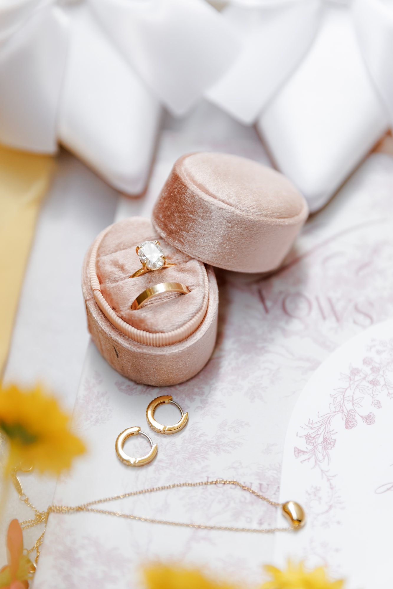 Gold engagement ring with diamond solitaire in pink velvet ring box, with hoop earrings and delicate necklace on vows paper.