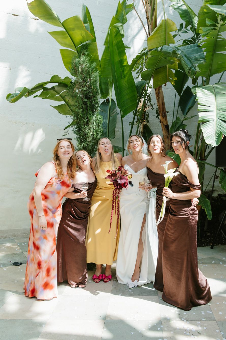 Bride and bridesmaids in colorful mismatched dresses posing playfully in front of tropical plants outdoors.