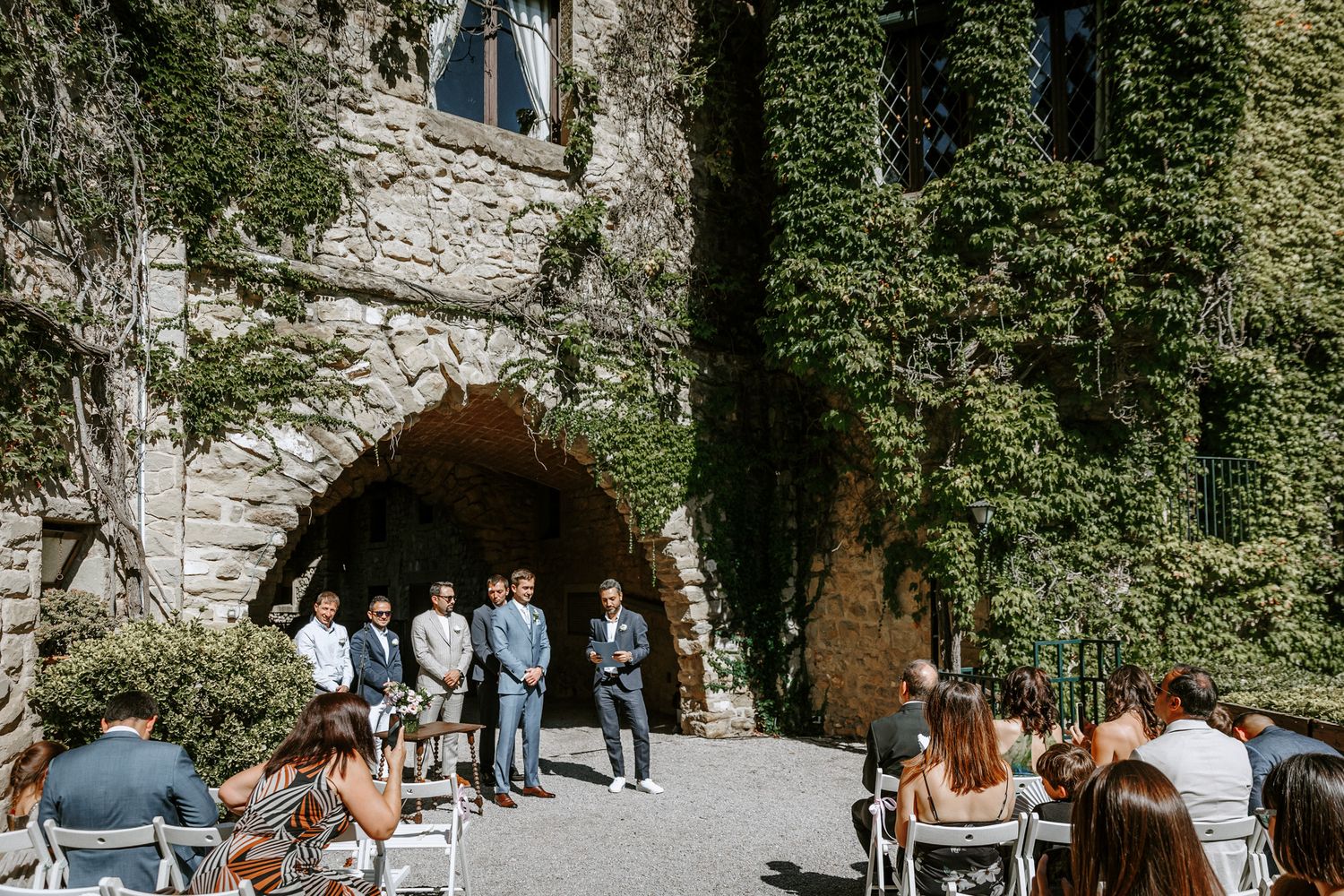 Outdoor wedding ceremony at La Baronia wedding venue with ivy-covered walls, groomsmen at the altar, guests seated in white chairs.