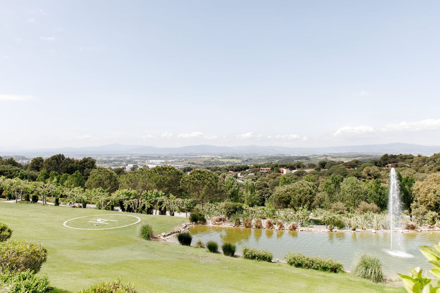 Panoramic view of lush green gardens with a fountain pond and sprawling landscape under a clear blue sky.