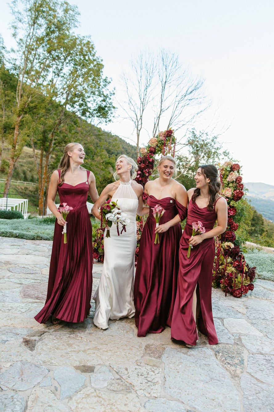 Bride in white gown laughs with three bridesmaids in burgundy satin dresses holding bouquets near a floral arch on a stone patio.