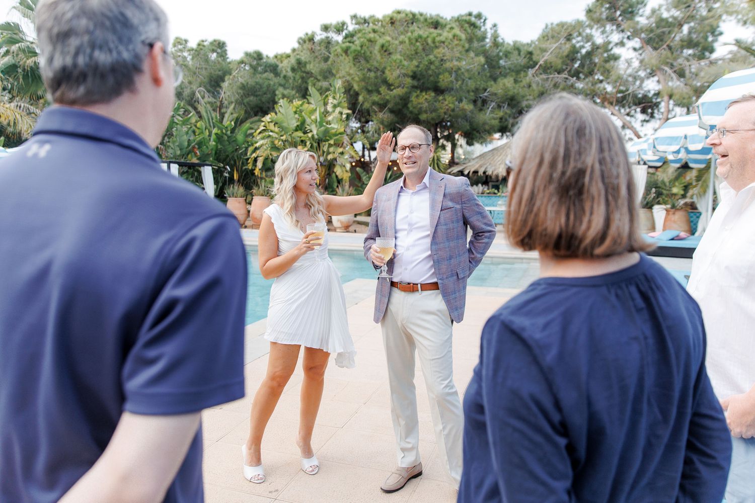 Group of adults socializing outdoors near a pool at a sunny resort, dressed in casual summer attire.