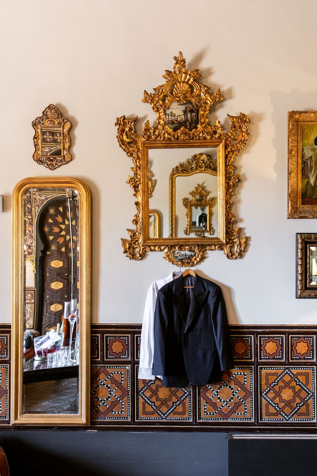 Ornate gold baroque mirror above a hanging suit jacket on a tiled wall with a tall floor mirror reflecting an elegant room.