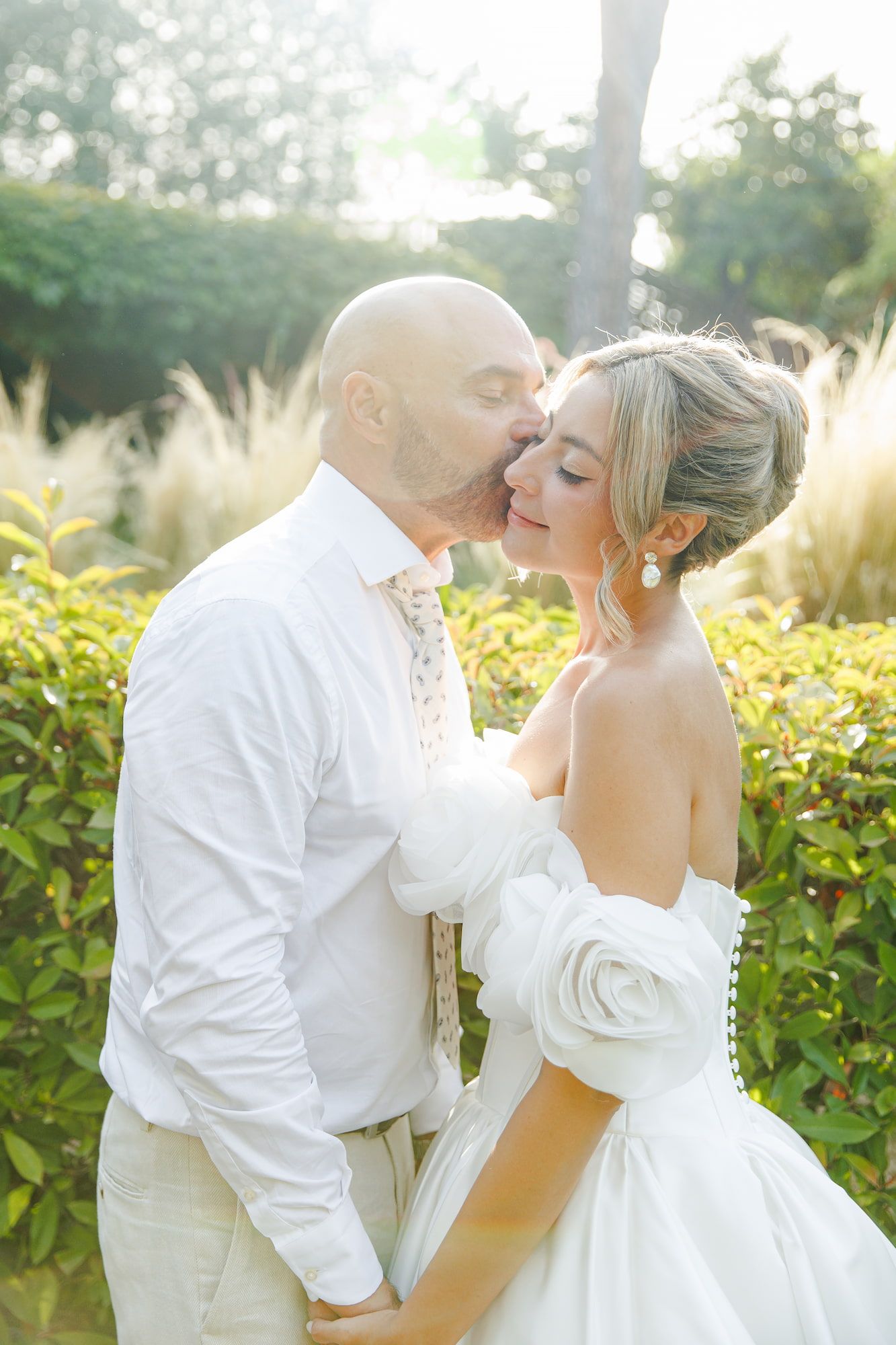 Groom kissing bride on cheek outdoors, both in white wedding attire, surrounded by lush green hedges.