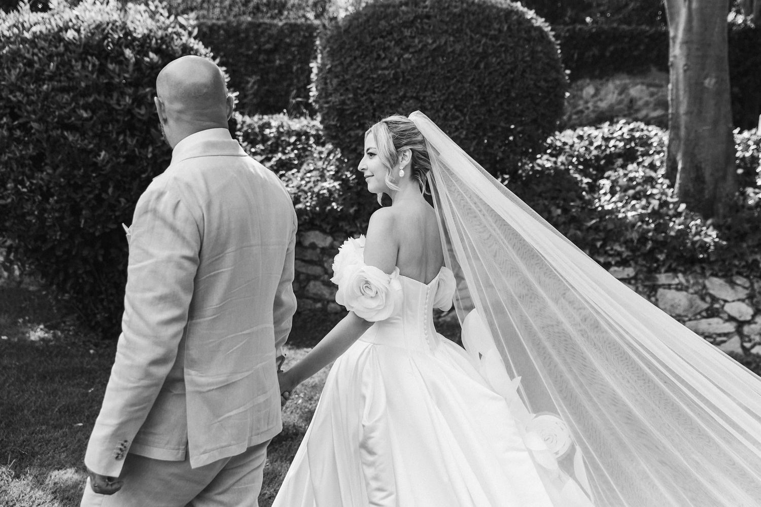 Black and white photo of a bride in a flowing gown and veil walking with a groom in a suit through a garden.