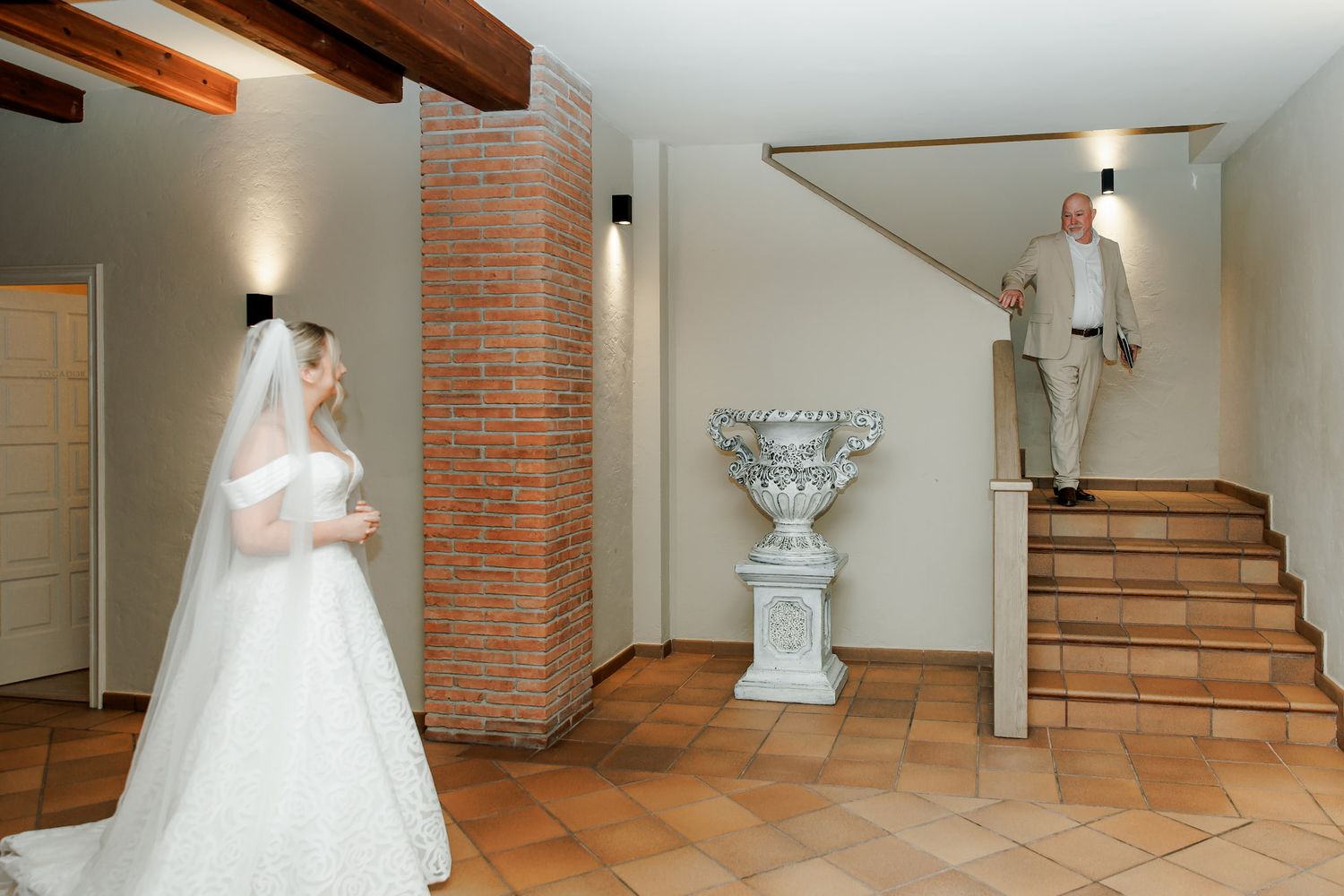 Bride in white gown and veil stands in elegant venue as man descends staircase near decorative urn.