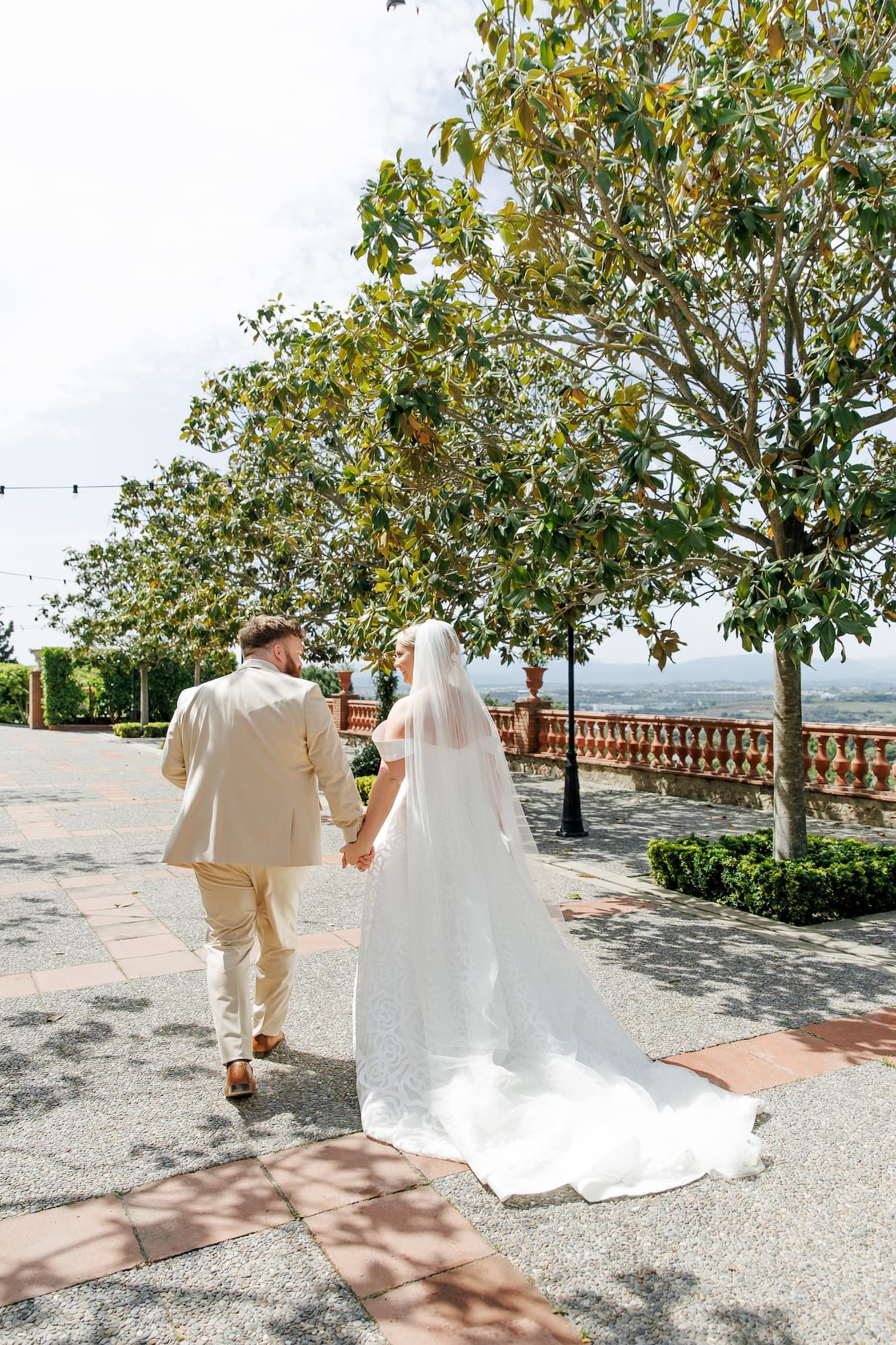 Bride and groom walk hand-in-hand along a sunny outdoor terrace with lush trees and a balustrade backdrop.