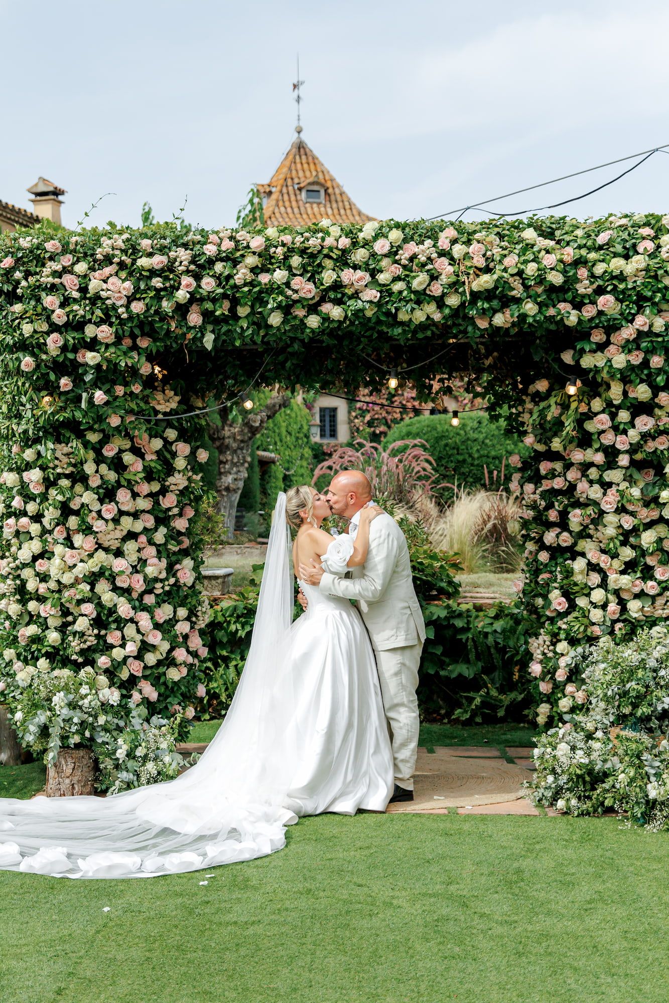 Bride and groom share a kiss under a floral arch adorned with pink roses at an outdoor wedding ceremony.