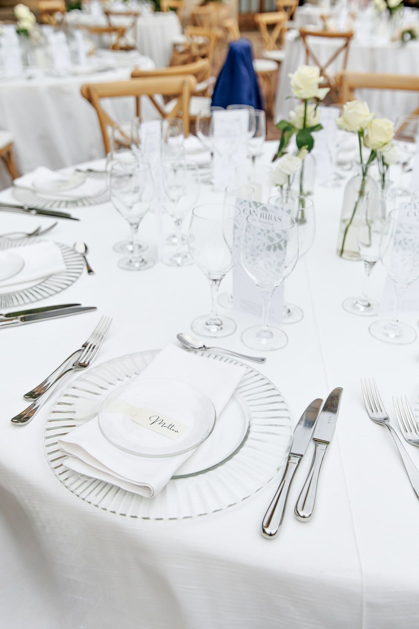 Elegant white wedding reception table setting with crystal glasses, silver cutlery, white roses, and folded napkins on white tablecloth.