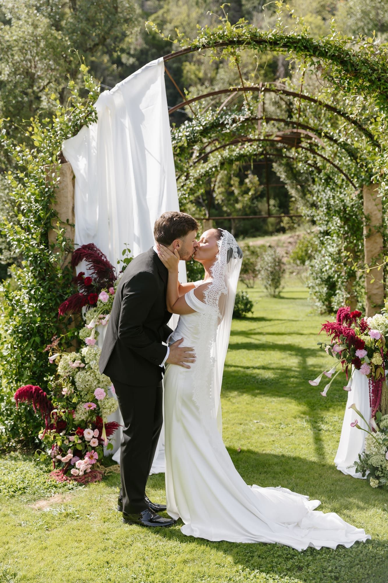Bride and groom share first kiss under floral arch with white draping and lush greenery at outdoor wedding ceremony.