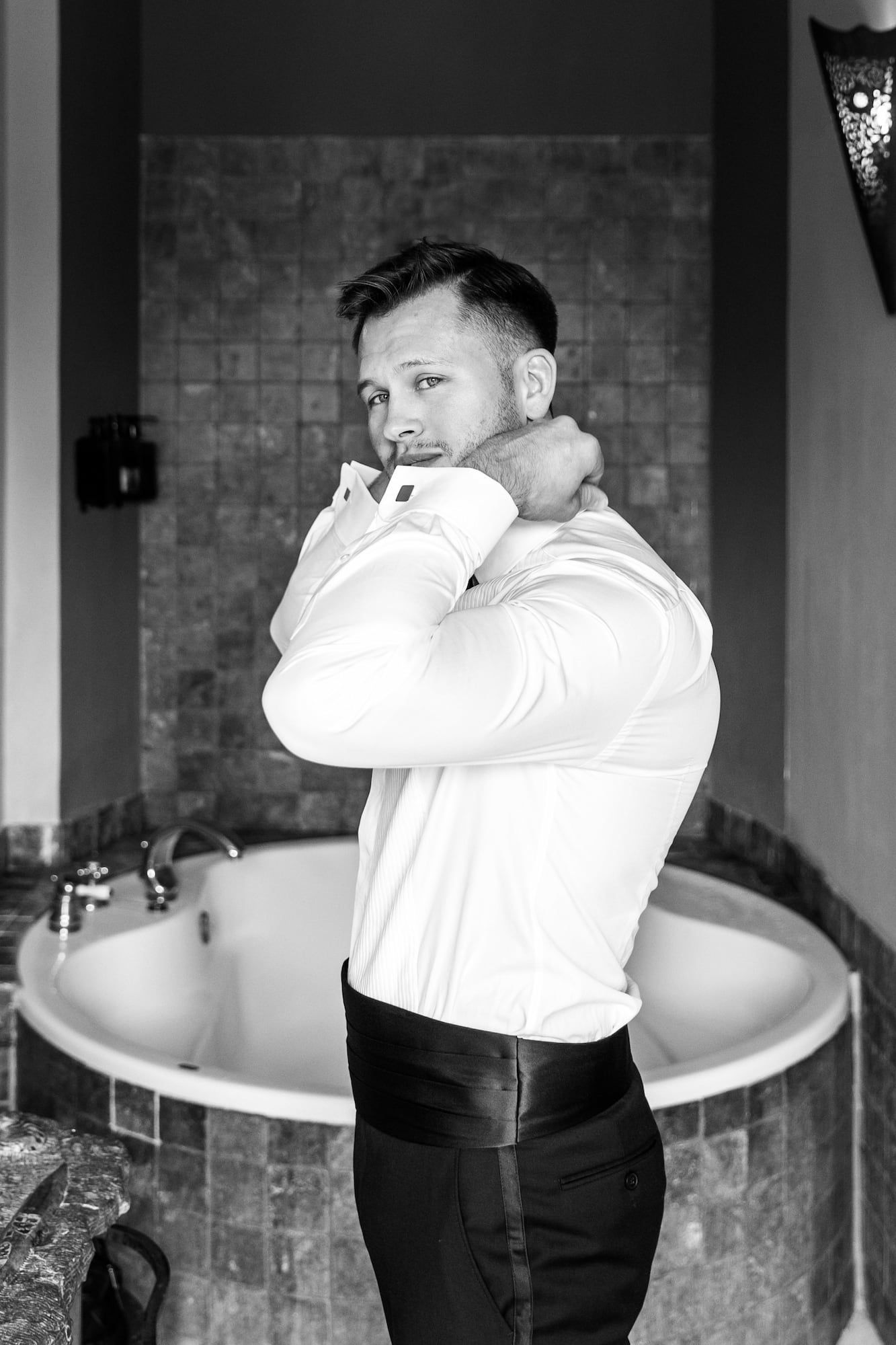 Black and white photo of a muscular man in white dress shirt adjusting his collar near an elegant bathroom tub.