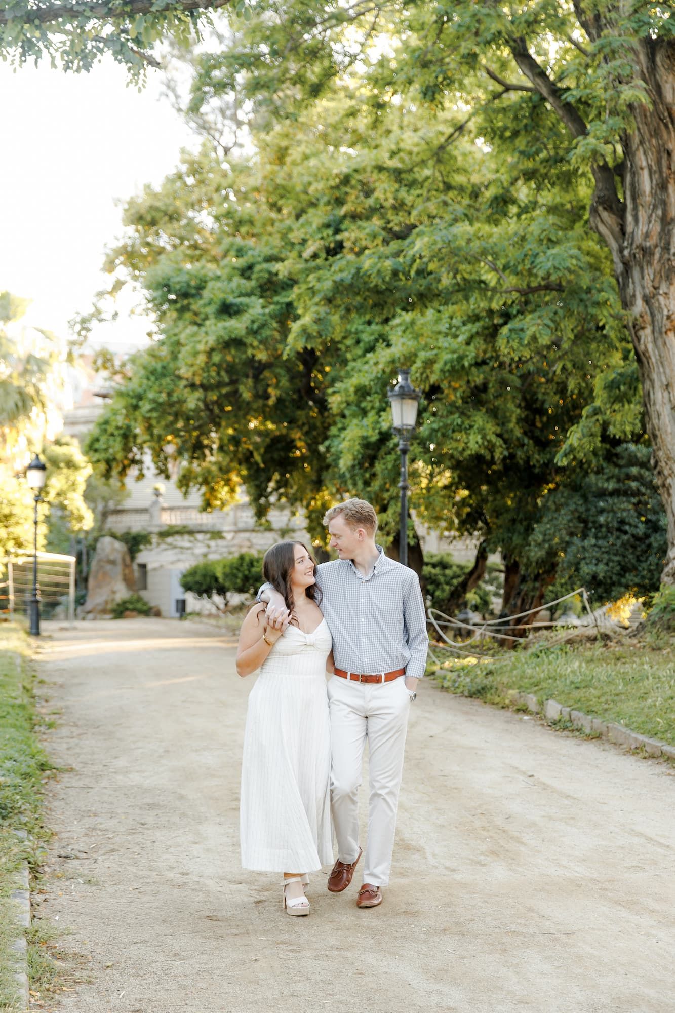 Couple walks hand-in-hand on a sunlit garden path, surrounded by lush green trees, woman in white dress, man in blue shirt.