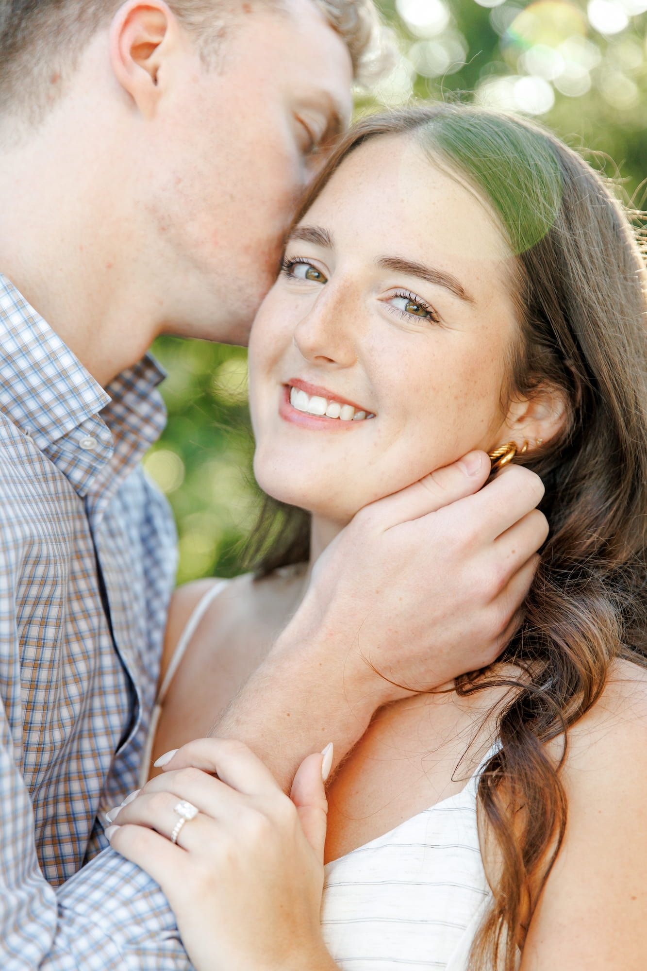 A man kisses a smiling woman's cheek while cradling her face, she wears an engagement ring and gold earring.