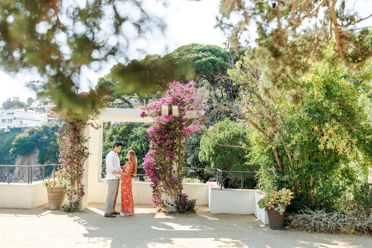 Couple standing together in a lush garden with pink bougainvillea and green trees on a sunny day.