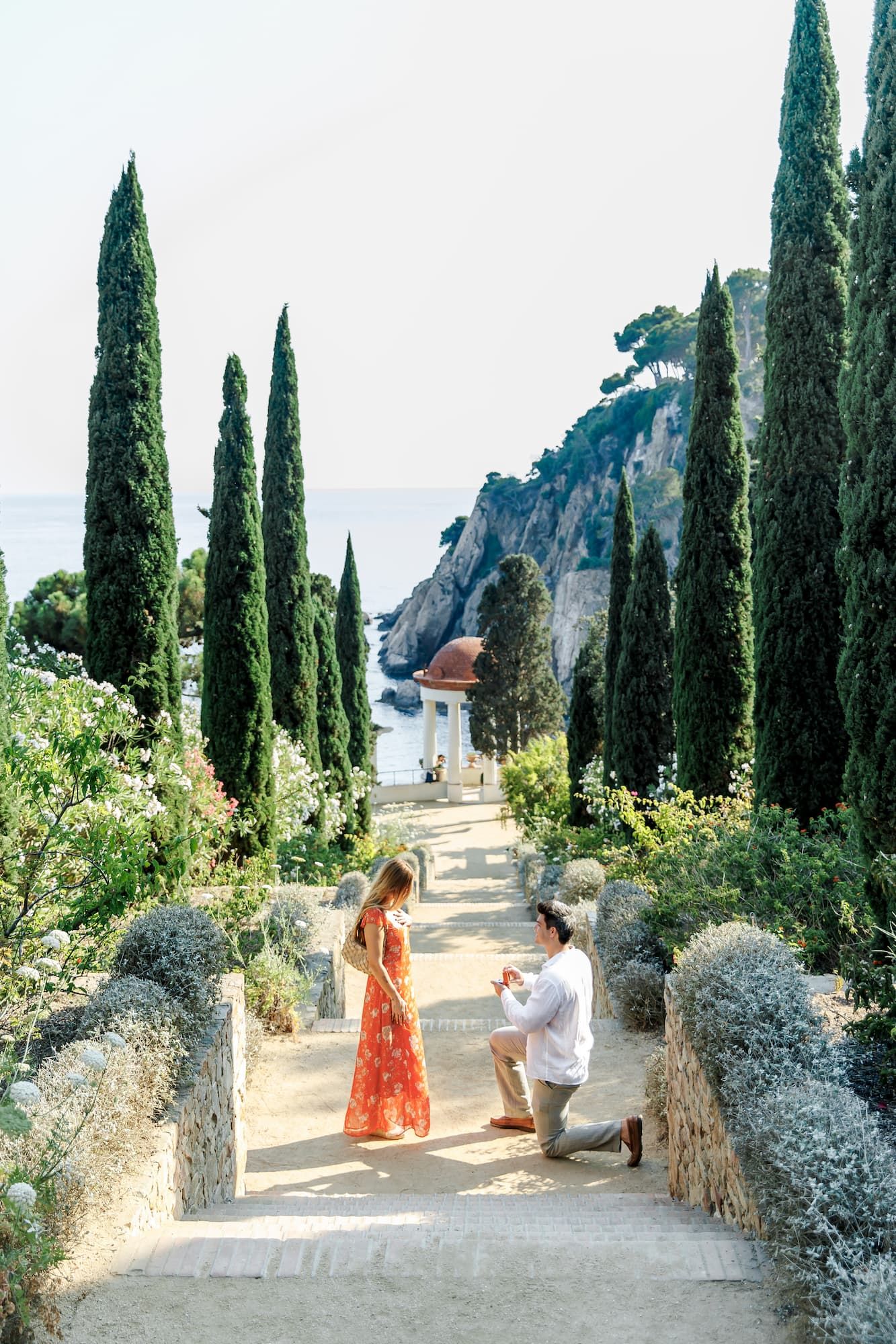 Man proposes on one knee in a stunning Italian garden with cypress trees and Mediterranean sea views.
