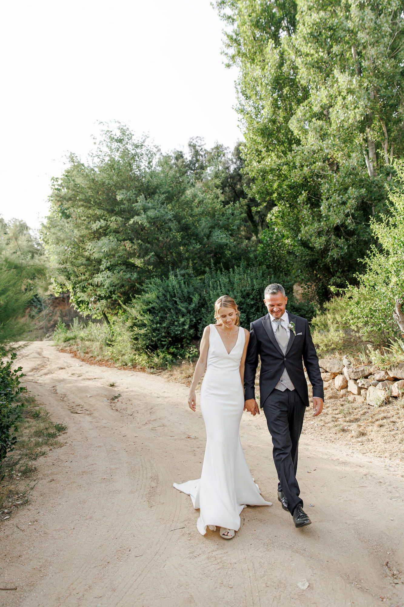 Bride in white gown and groom in suit walk hand in hand on a sandy path surrounded by lush green trees.