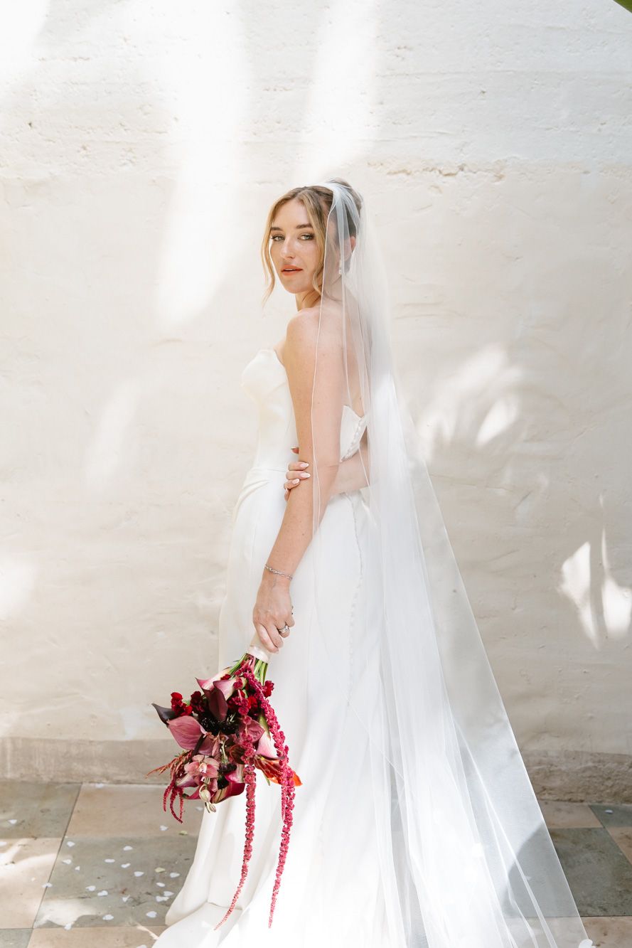 Elegant bride in backless white gown and veil holds dark red bouquet against sunlit white wall at Masia Victoria
