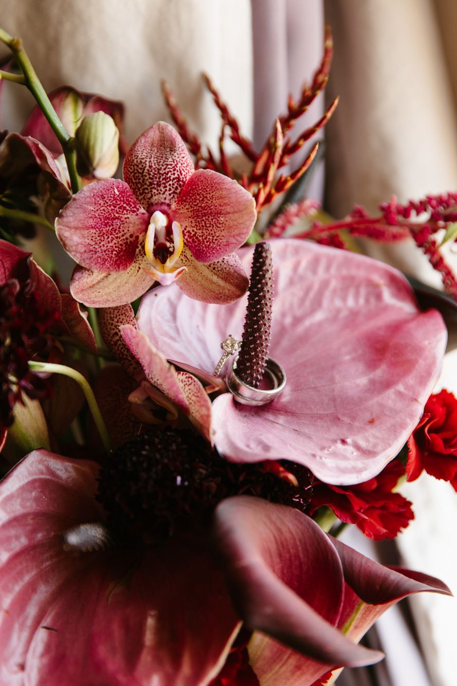 Elegant wedding rings resting on a pink anthurium surrounded by orchids and red flowers in a bridal bouquet.