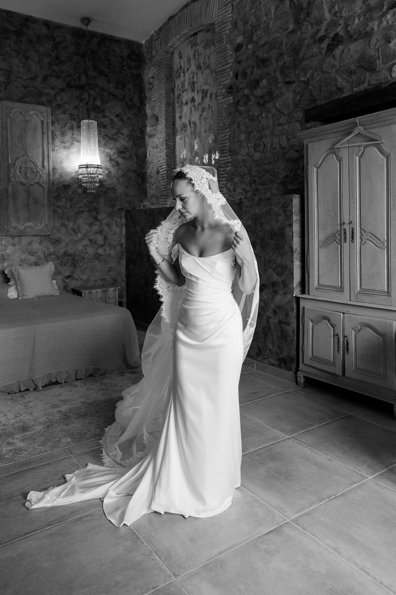 Elegant bride in a white gown and lace veil poses in a rustic stone room in this black and white photo.