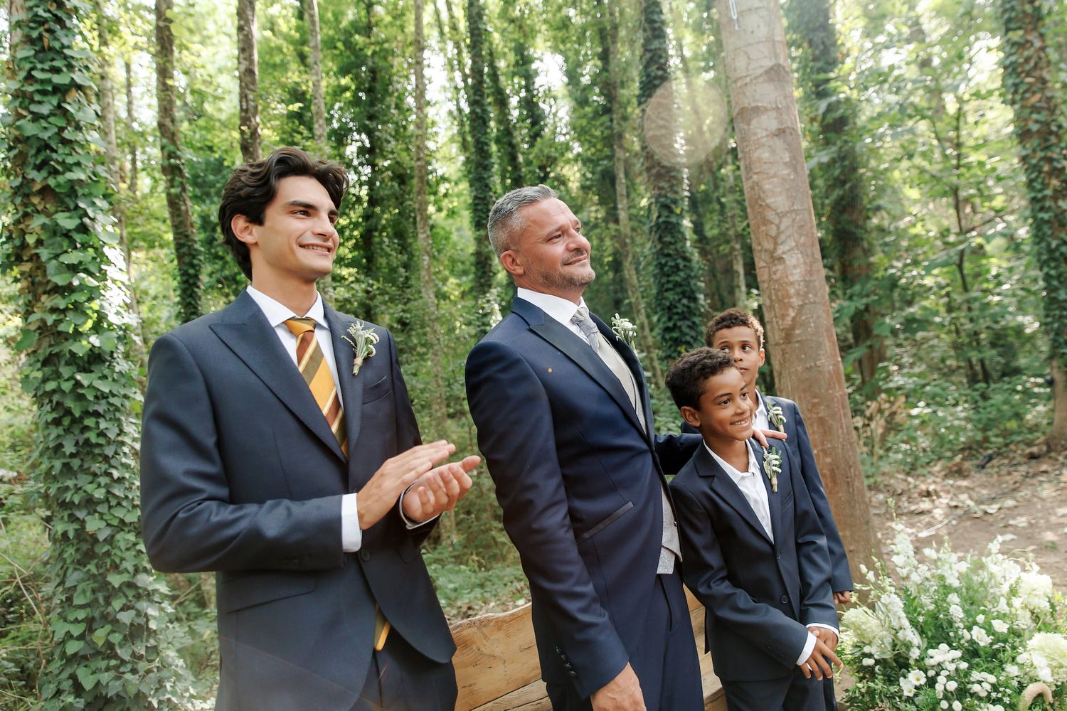 Three men in navy suits smile outdoors at a woodland wedding ceremony surrounded by lush green ivy.