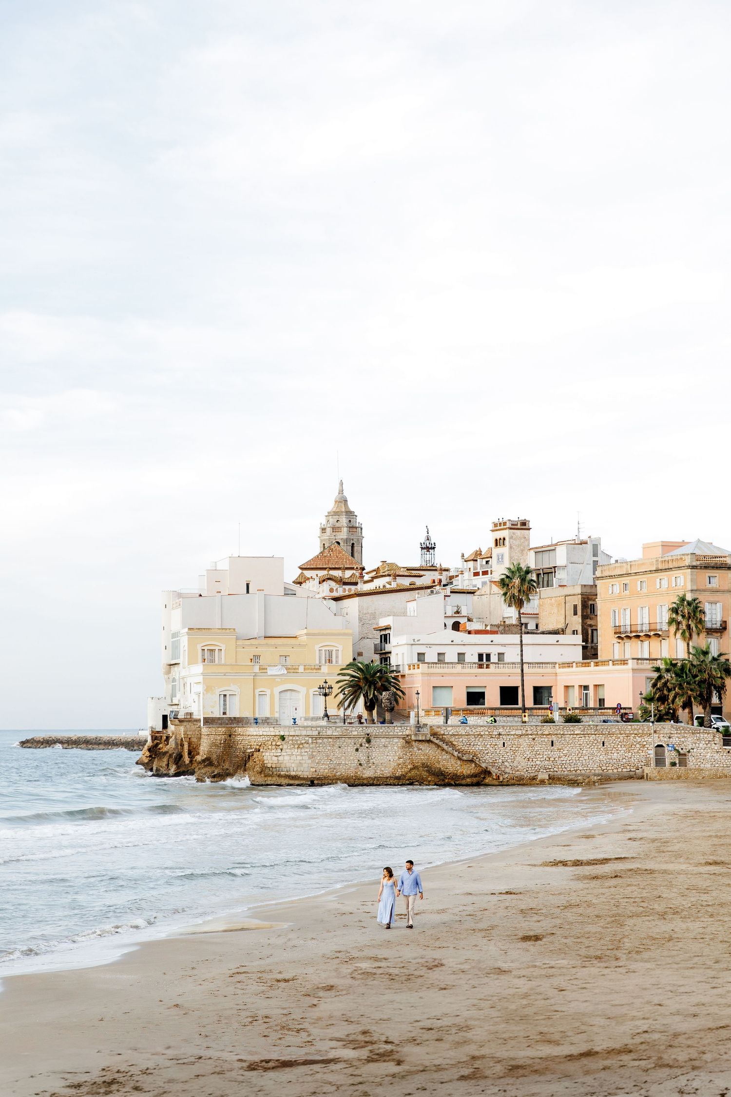 An engaged couple walks along the coastline, laughing, with the city of Sitges in the background.