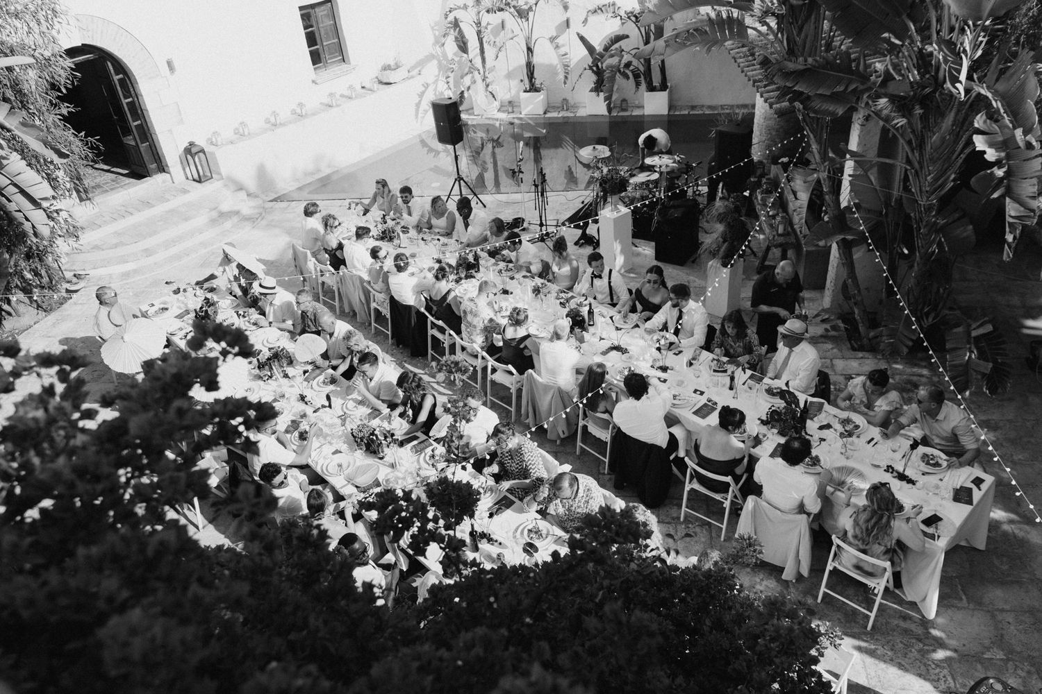 Aerial black and white photo of a large outdoor dinner party with guests seated at long tables in a courtyard.
